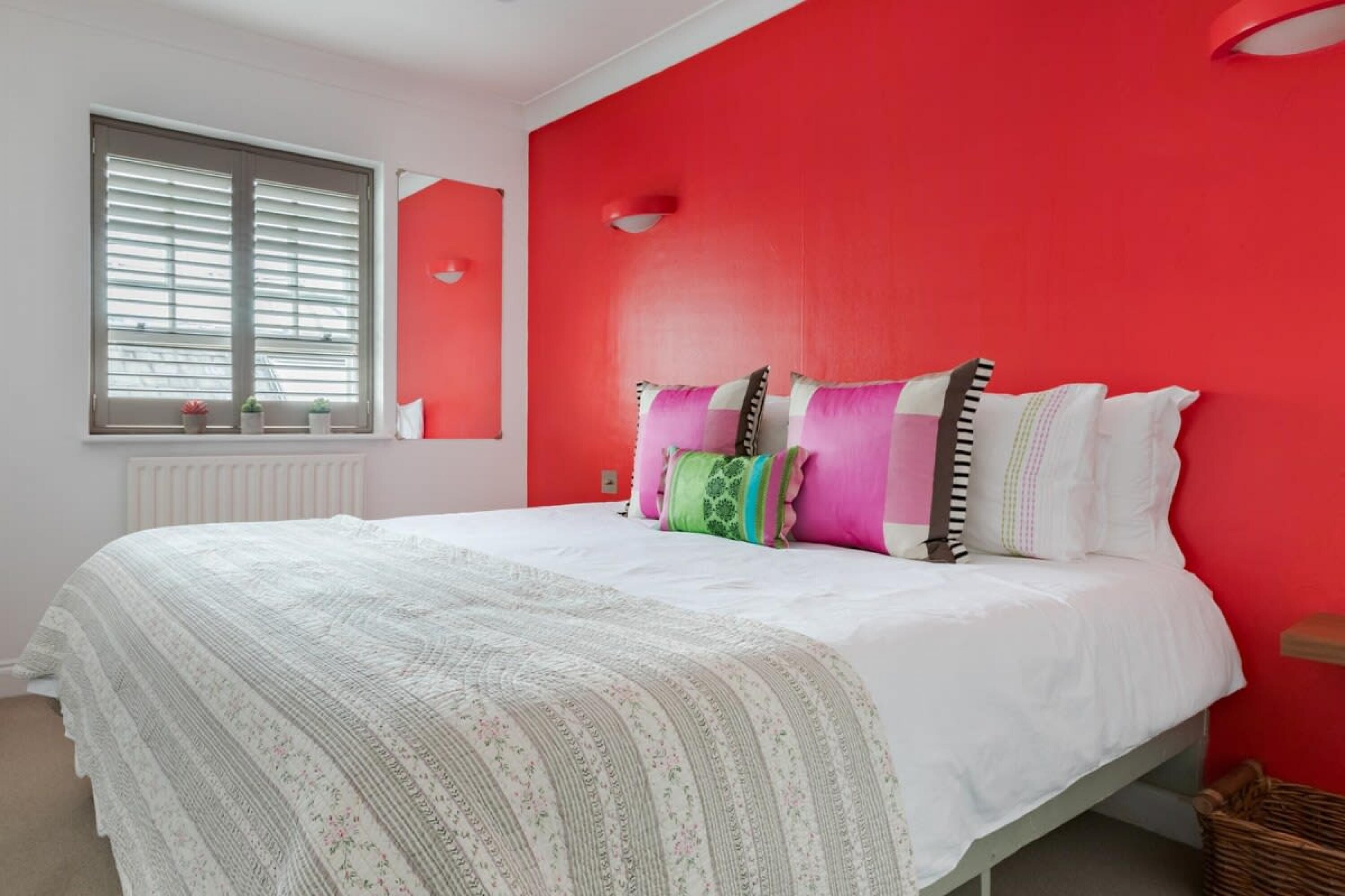 Une chambre à coucher avec un lit blanc, des murs rouges et une fenêtre avec des stores.