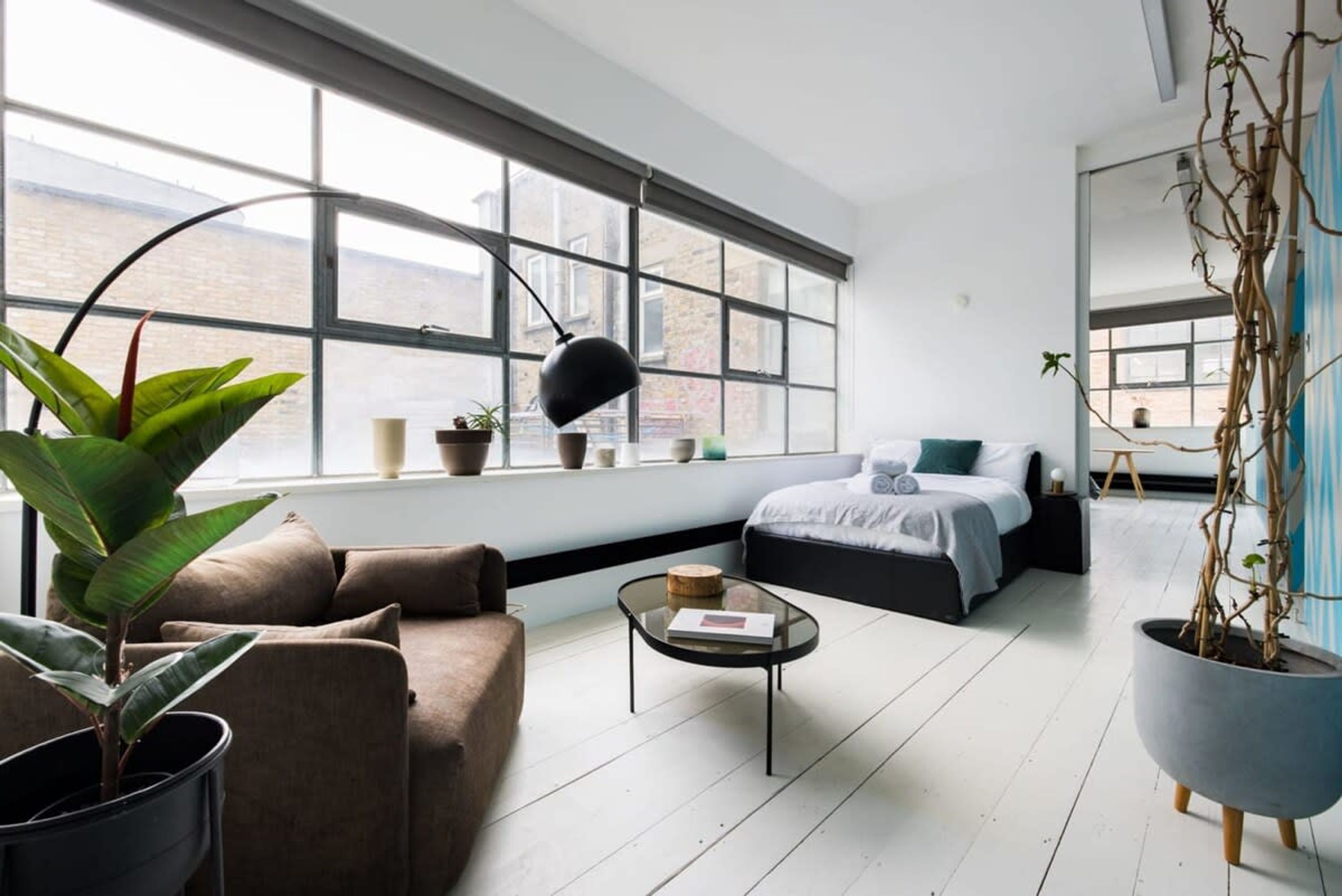 A modern living room with a large window, a sofa, a coffee table, and a bed with a blue accent pillow.