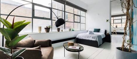 A modern living room with a large window, a sofa, a coffee table, and a bed with a blue accent pillow.