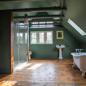 A spacious bathroom with a wooden floor, a glass shower enclosure, a white sink, and a clawfoot bathtub.
