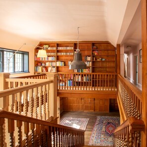 Une bibliothèque spacieuse avec des étagères en bois, un grand lustre suspendu et un escalier en bois surmonté d’un tapis à motifs.