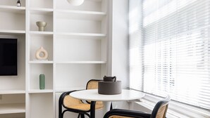 A modern room with a white wall, a flat-screen TV mounted on the wall, a round table with chairs, and a window with blinds. - Light-filled Studio With AC - Earl s Court (London)