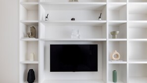 A modern living room with a flat-screen TV mounted on the wall, surrounded by white shelving units with various decorative items. - Light-filled Studio With AC - Earl s Court (London)
