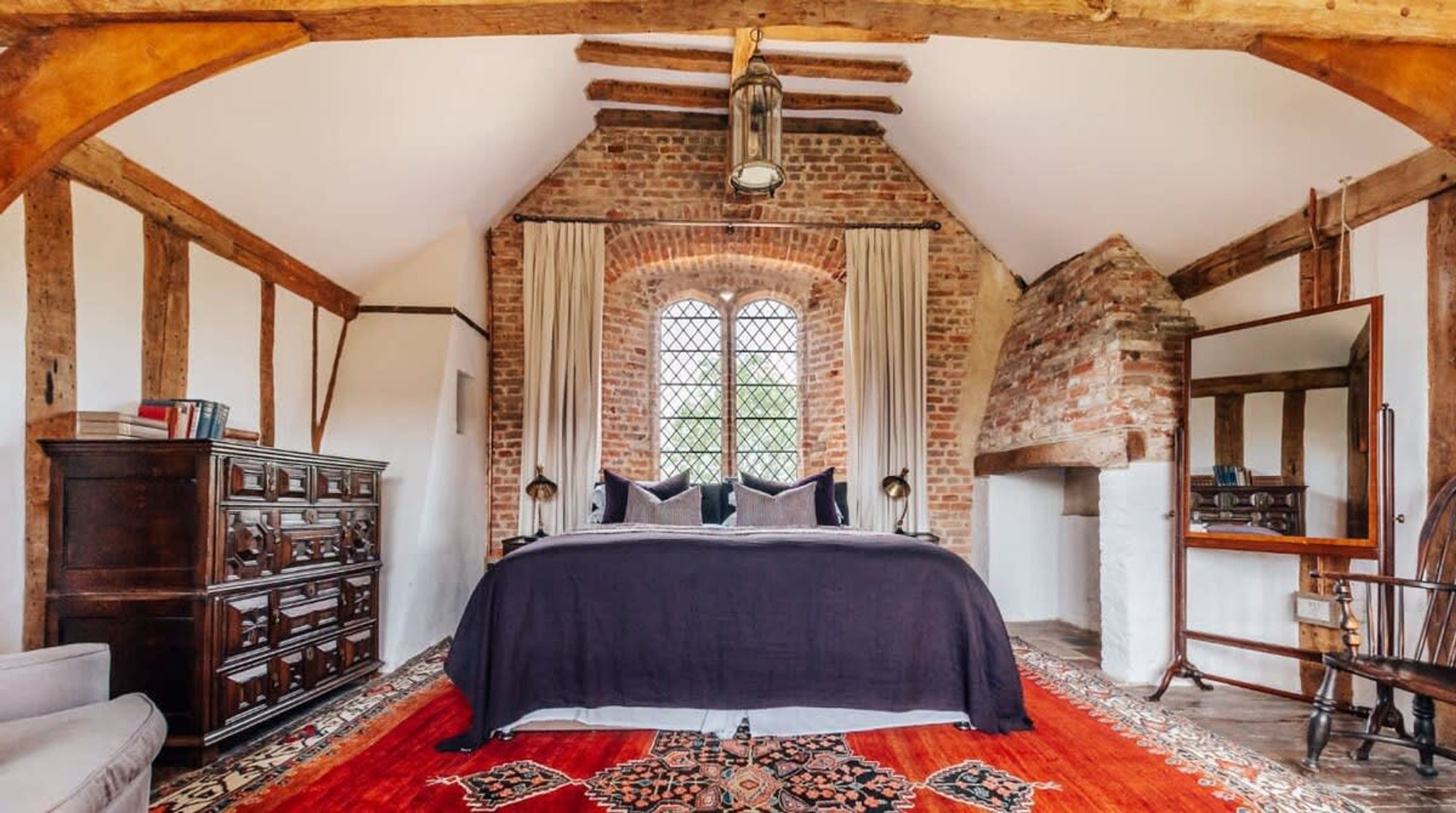 A bedroom with a large bed, a wooden dresser, a red patterned rug, and a brick wall with a window.