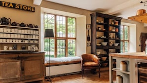 Une pièce chaleureuse avec une grande fenêtre, une bibliothèque remplie d’objets liés au thé et un fauteuil placé près de la fenêtre.