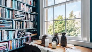 Een kamer met een grote boekenkast vol boeken, een raam met uitzicht op bomen, en een tafel met kaarsen en aardewerk.