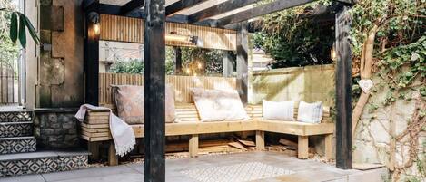 A wooden pergola with a bench and cushions, stone steps, and a stone wall with ivy.