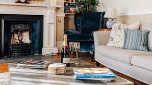 Een gezellige woonkamer met een open haard, een comfortabele fauteuil, een bank en een bijzettafel met boeken en een fles champagne.