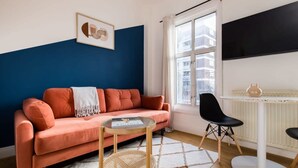 Ein Wohnzimmer mit blauer Akzentwand, einem rosafarbenen Sofa, einem runden Couchtisch, einem schwarzen Sessel und einem weißen Heizkörper.