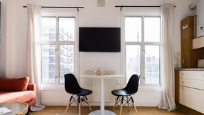Ein modernes Wohnzimmer mit einer Couch, einem runden Tisch mit zwei Stühlen, einem an der Wand montierten Fernseher und großen Fenstern mit Vorhängen.