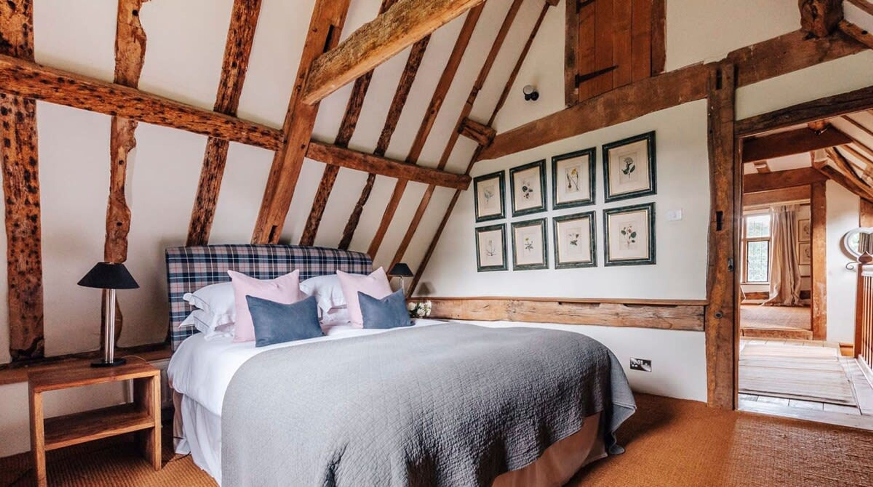 A bedroom with a wooden ceiling, a bed with a plaid headboard, and framed botanical prints on the wall.