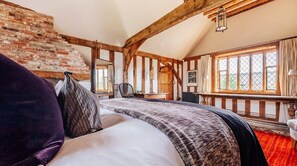 Een slaapkamer met zichtbare houten balken, een bakstenen muur, een groot raam met gordijnen en een bed met een beddegoed met patroon.