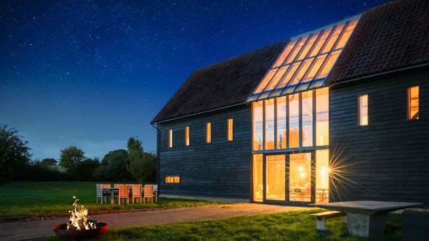 A wooden building with large windows and a fire pit in the foreground. - Grand Suffolk Barn Conversion (Ipswich)