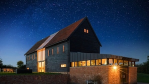 A modern house with a sloped roof and large windows, set against a night sky with stars. - Grand Suffolk Barn Conversion (Ipswich)