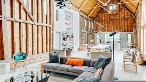 A spacious living area with a wooden ceiling, a grey sofa with orange cushions, and a dining area in the background. - Grand Suffolk Barn Conversion (Ipswich)