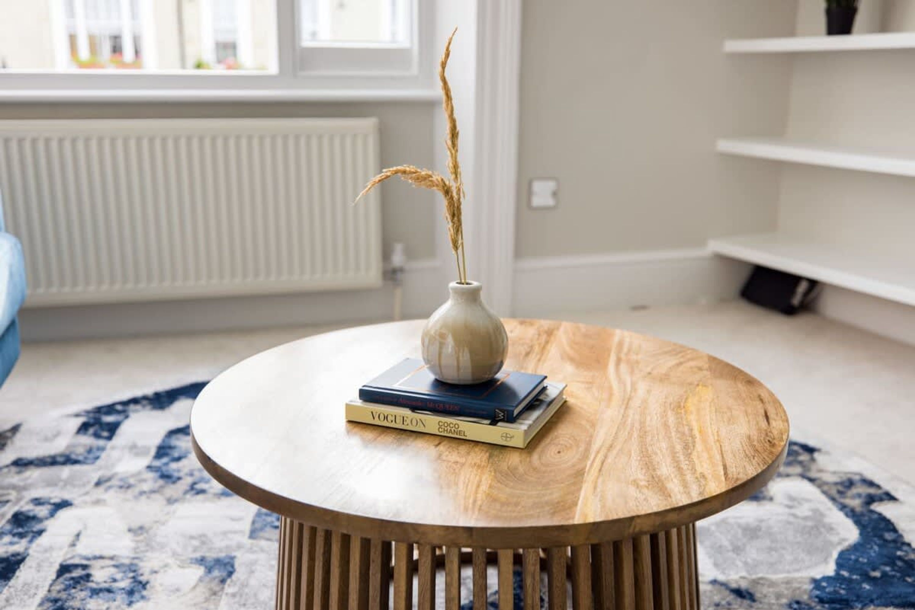 Ein runder Holztisch mit einer weißen Vase und Büchern, einem blauen Teppich mit Muster sowie einem Fenster mit Blick ins Freie.