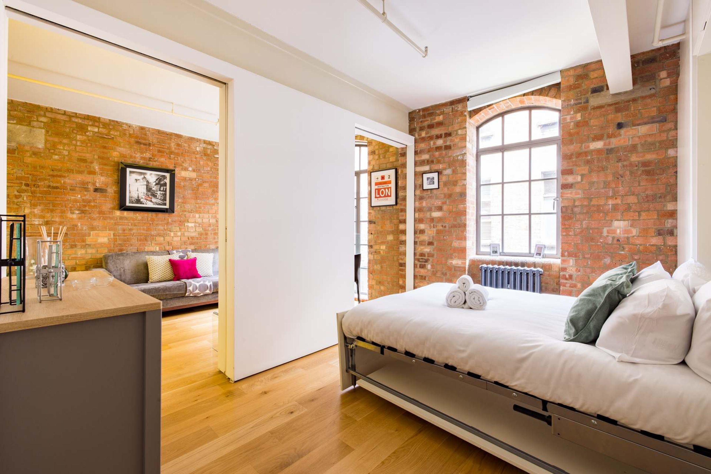 A modern bedroom with a brick wall, a large window, a bed with white bedding, and a grey sofa in the adjacent room.
