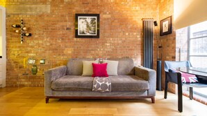 A living room with a brick wall, a grey sofa with patterned cushions, a wooden coffee table, and a window with a view. - Central Soho Loft With A C Concierge (London)