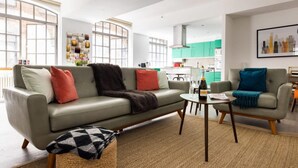 Un salon moderne avec un canapé gris, une table basse et une petite table d’appoint. On aperçoit à l’arrière-plan un coin cuisine avec des meubles verts et une table à manger.