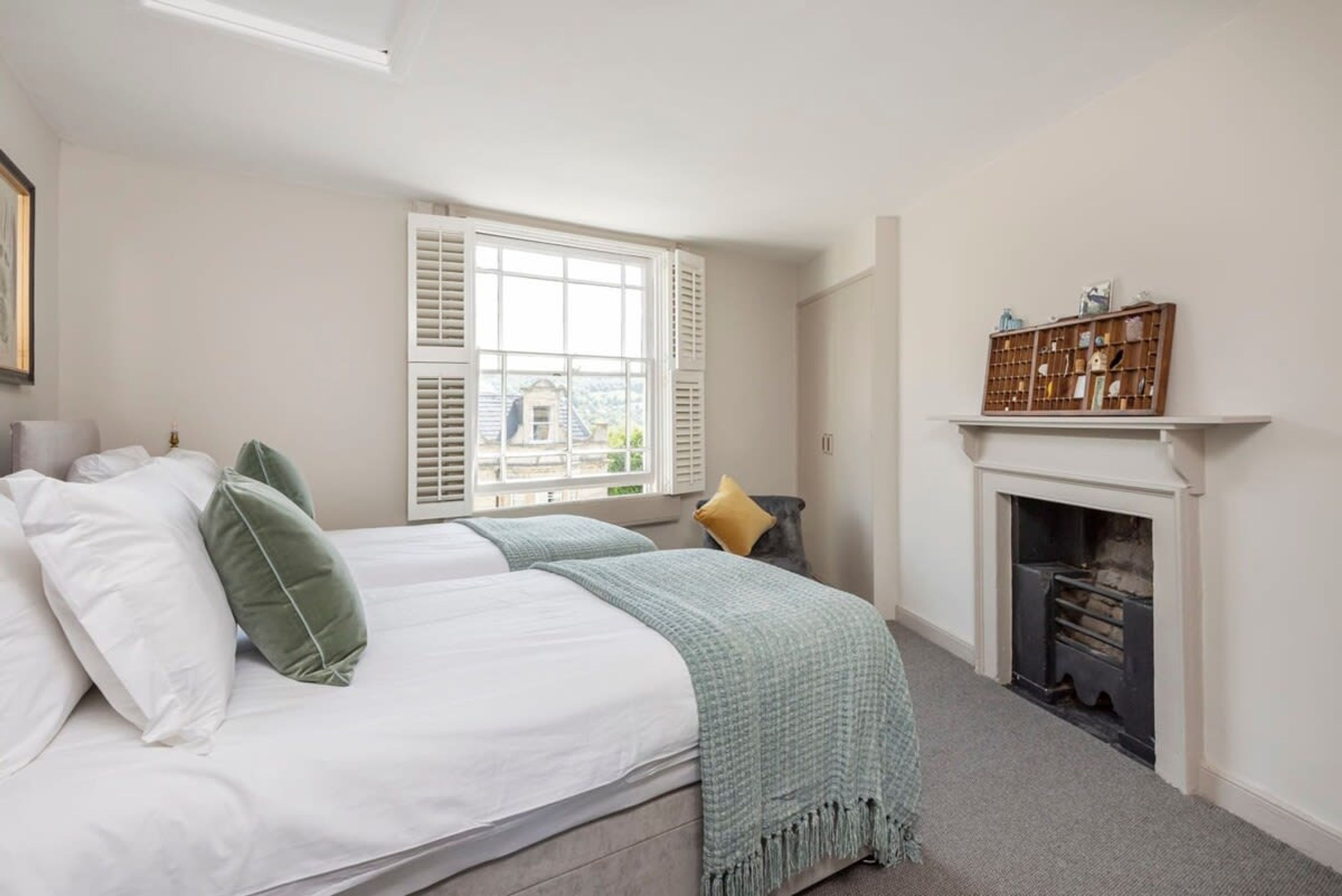 A bedroom with a bed, pillows, a window with shutters, a fireplace, and a shelf with decorative items.