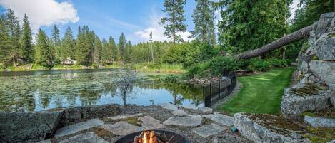 A lakeside outdoor area with a fire pit, surrounded by rocks and greenery.