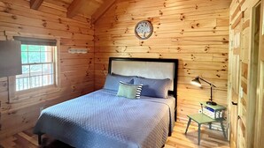 Quarto de cabana de madeira com cama, mesa de cabeceira, abajur e janela.