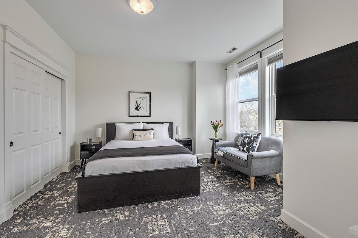 A bedroom with a bed, bedside tables, a chair, a window, and a wall-mounted TV.