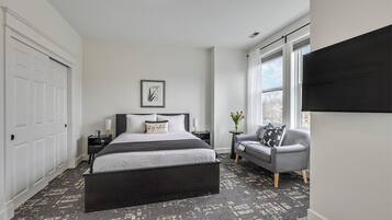 A bedroom with a bed, bedside tables, a chair, a window, and a wall-mounted TV.