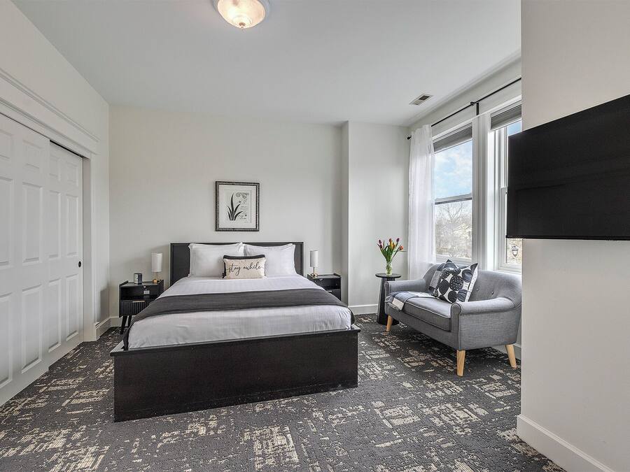 A bedroom with a bed, bedside tables, a chair, a window, and a wall-mounted TV.