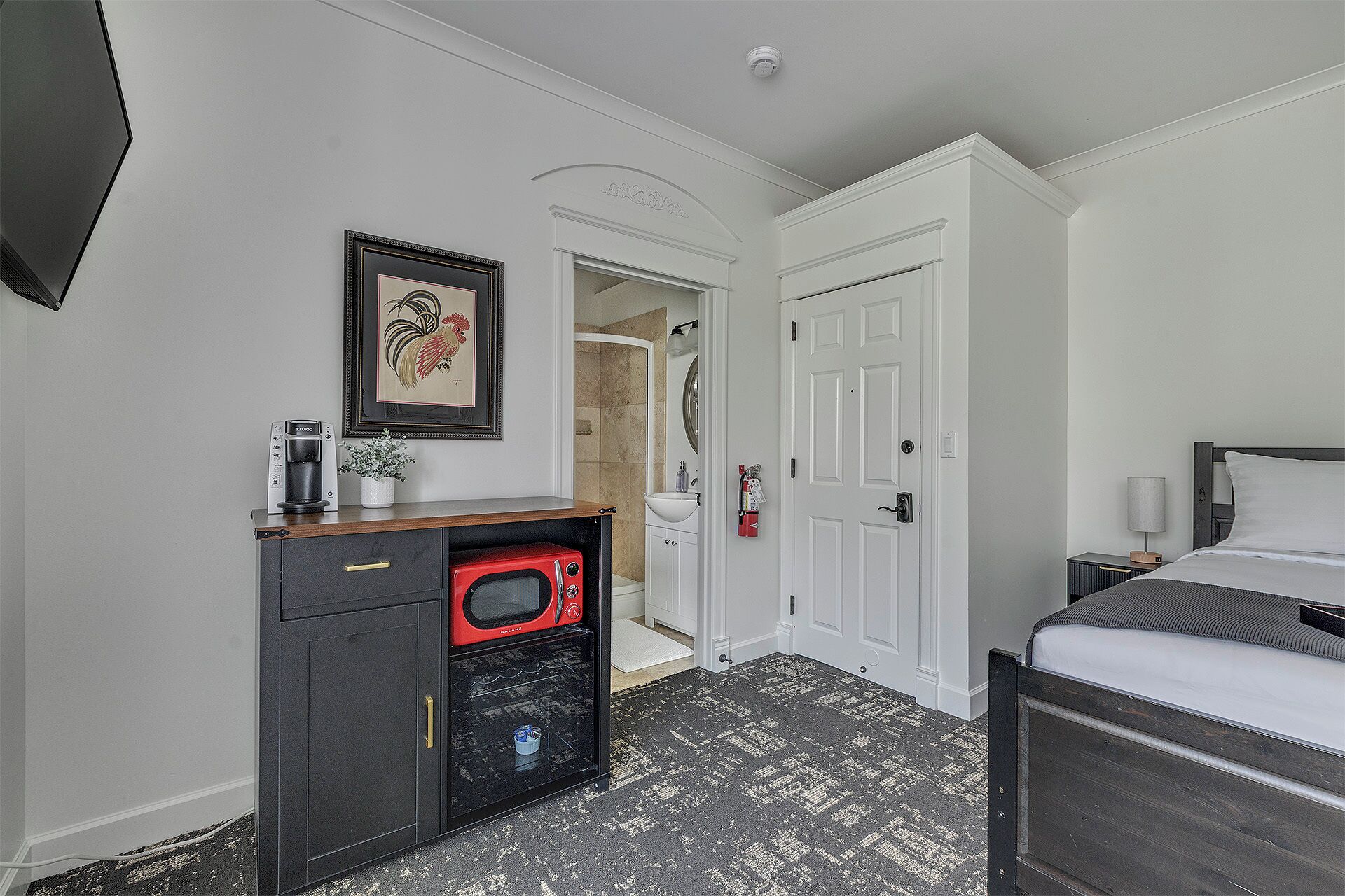 Hotelkamer met een bed, een bureau met een magnetron, een koffiezetapparaat, een inlijsting afbeelding, een witte deur en een zichtbare badkamer via een geopende deur.