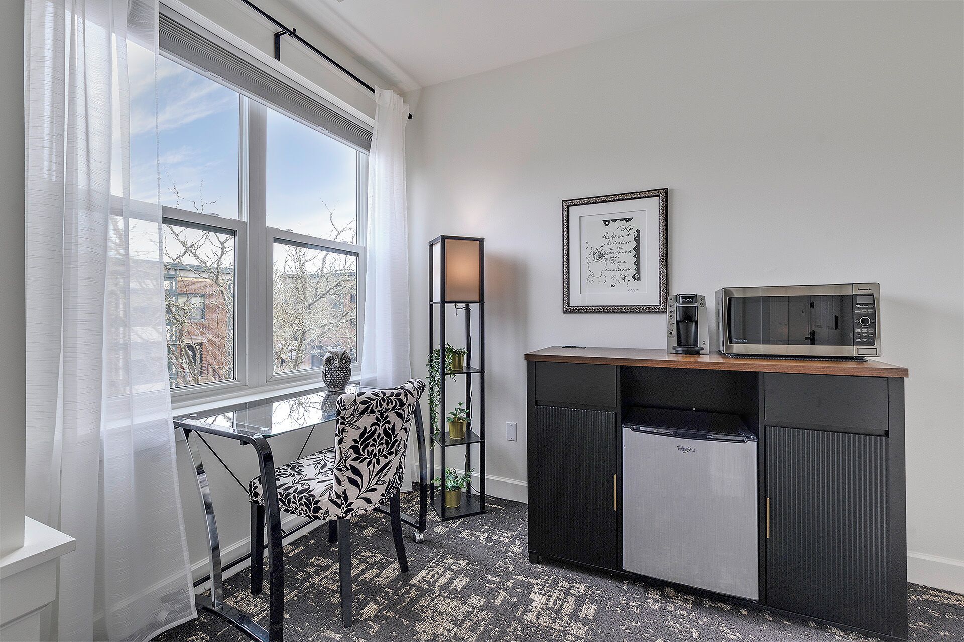 A compact kitchenette with a microwave, coffee maker, and refrigerator. There is a chair and a small table with a plant.