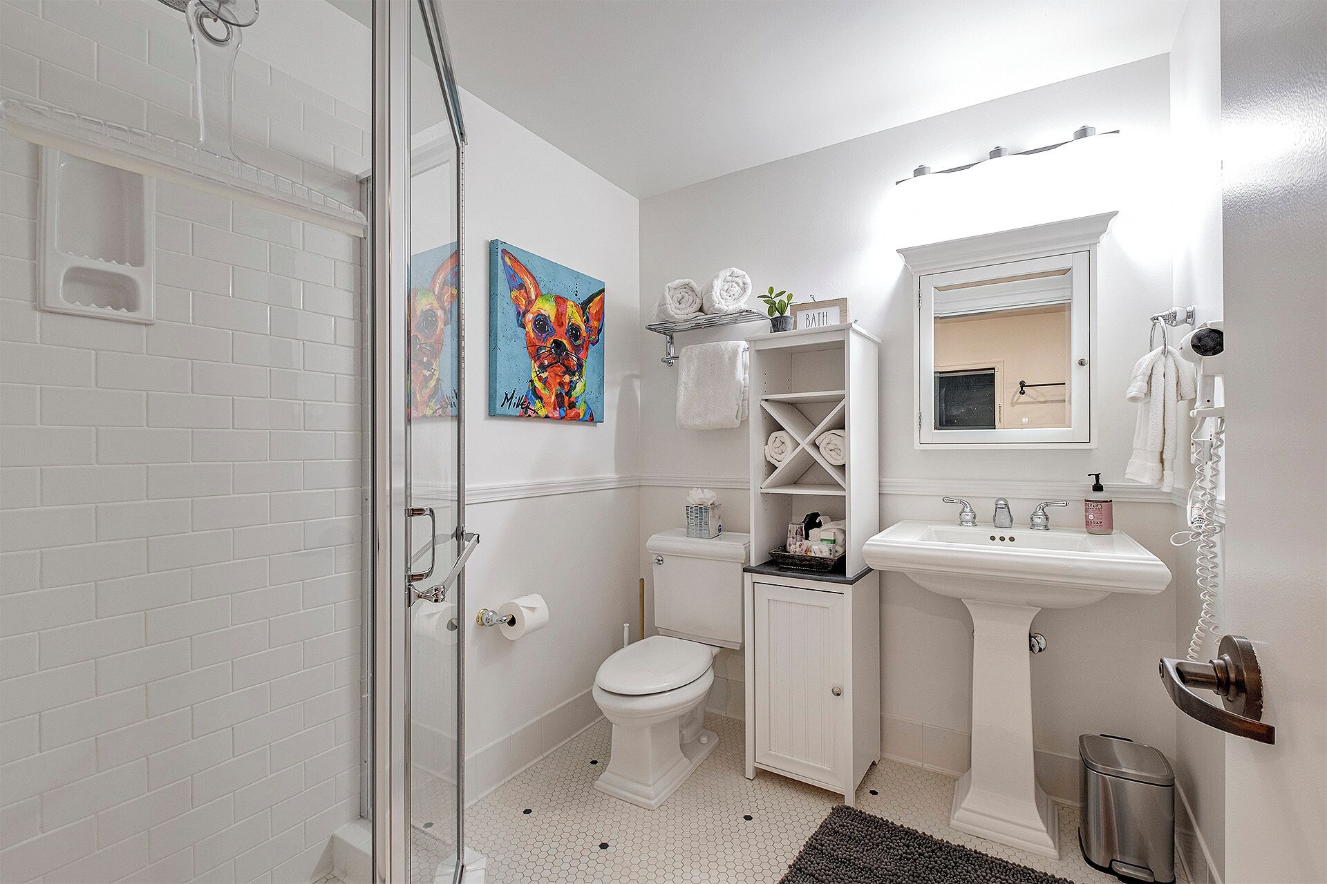 A bathroom with a glass shower enclosure, a toilet, a pedestal sink, and a small shelf with toiletries.