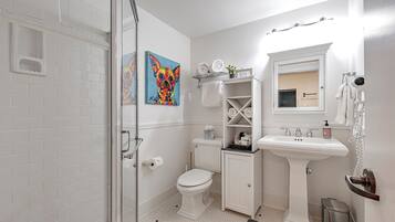 A bathroom with a glass shower enclosure, a toilet, a pedestal sink, and a small shelf with toiletries.