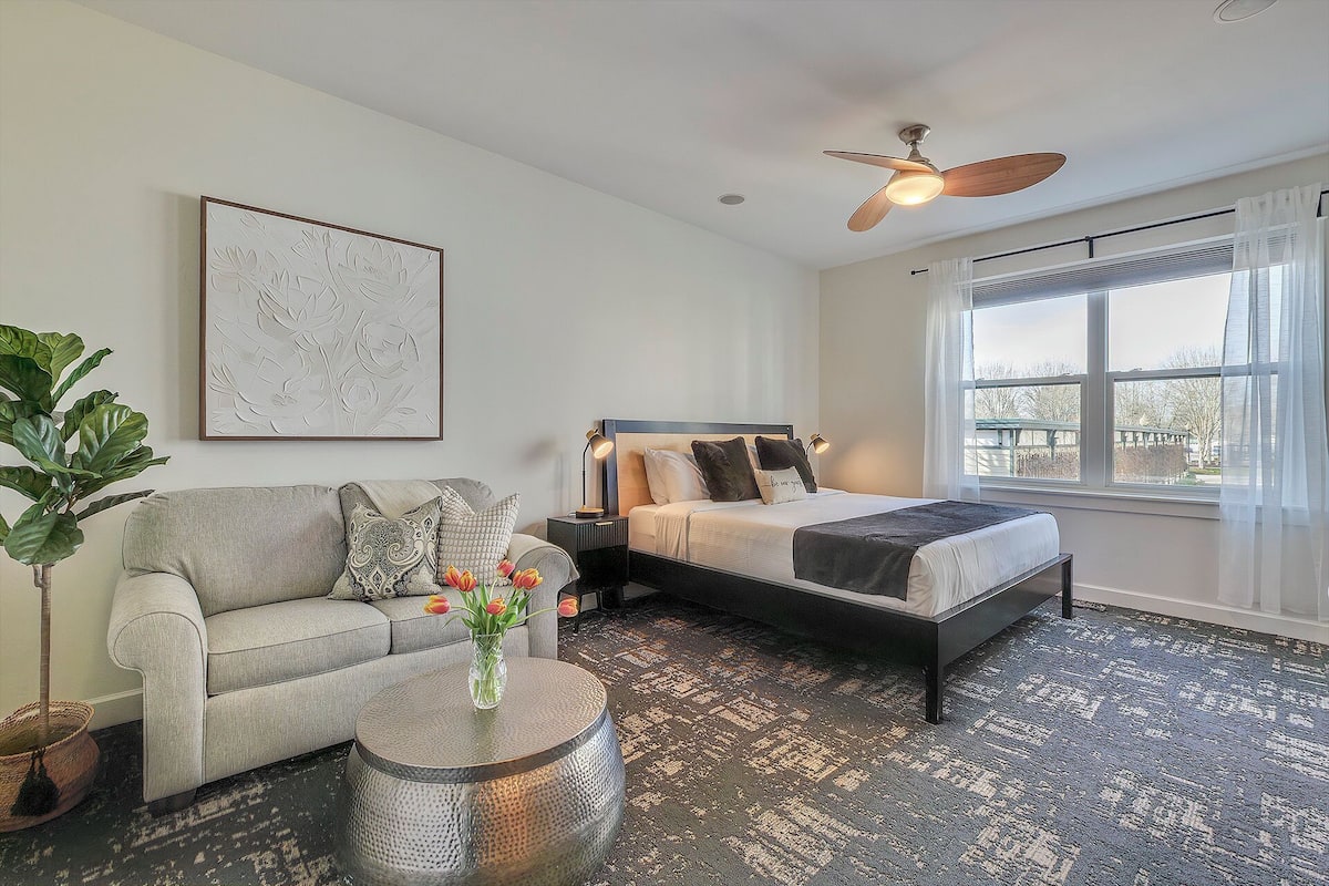 A bedroom with a bed, a sofa, a coffee table, a ceiling fan, and a window with curtains.