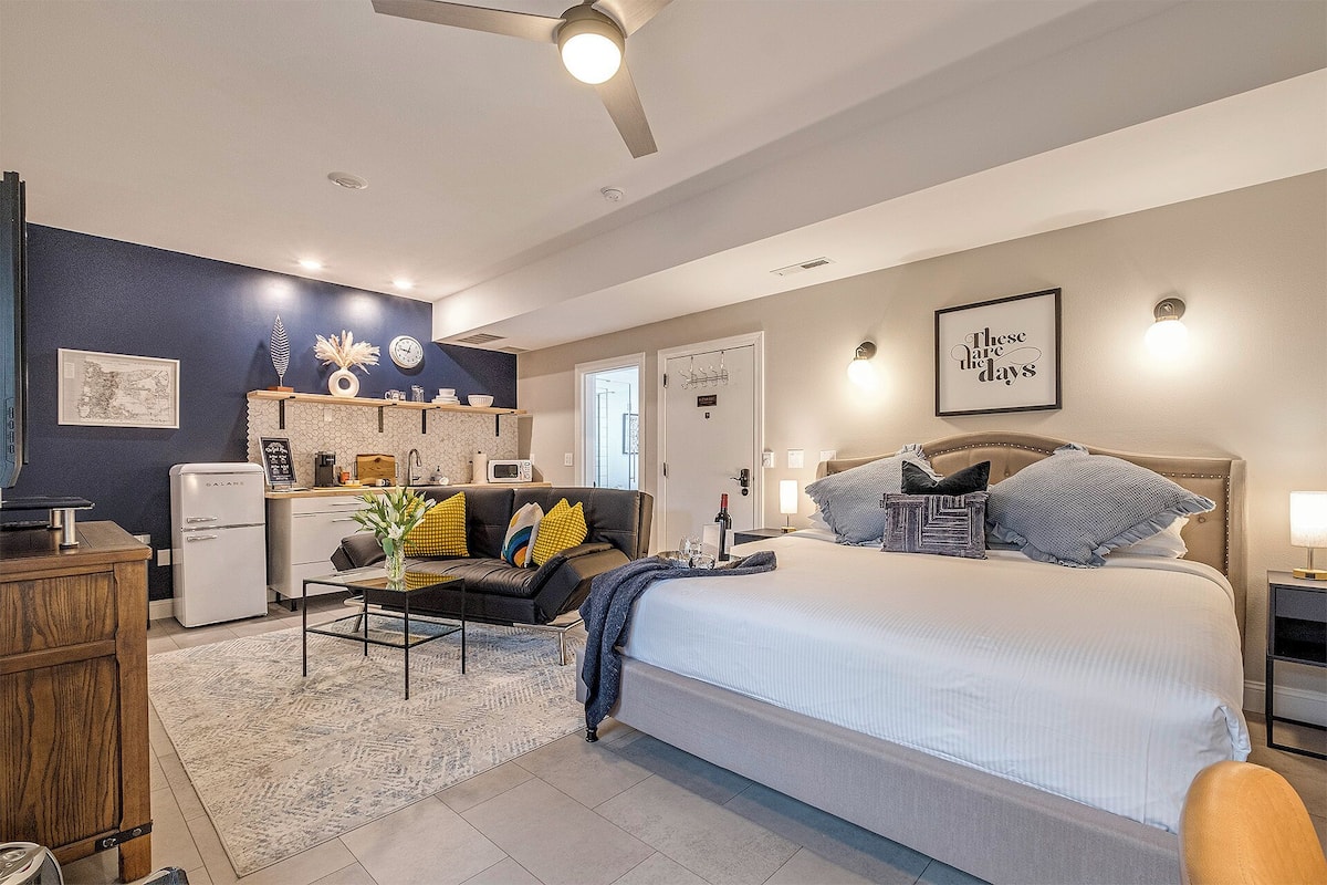A modern hotel room with a bed, a desk, a kitchenette, and a bathroom.