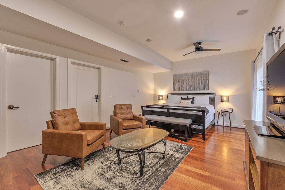 A spacious living room with a large bed, two leather armchairs, a glass coffee table, and a ceiling fan.