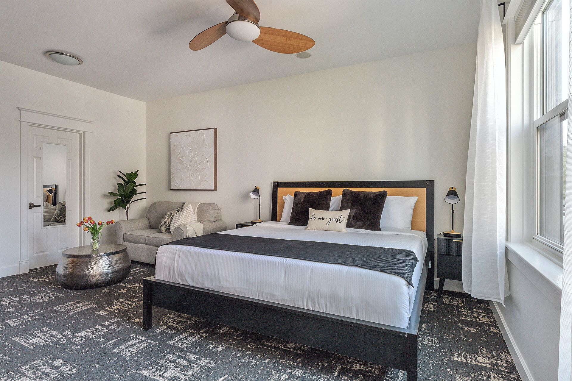 Une chambre à coucher comprenant un lit, un ventilateur de plafond, une table de chevet avec une lampe, une fenêtre avec des rideaux et une plante.