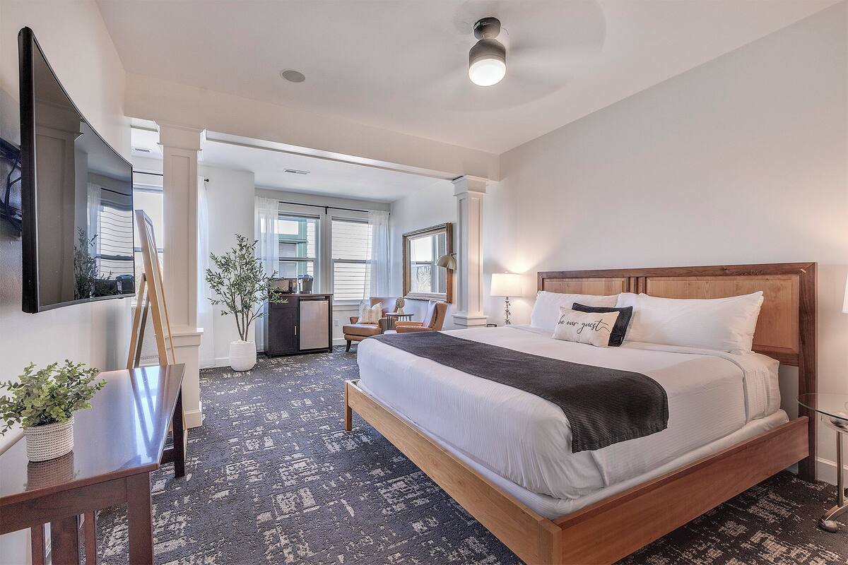 A hotel room with a large bed, a desk, a chair, a potted plant, and a mirror.