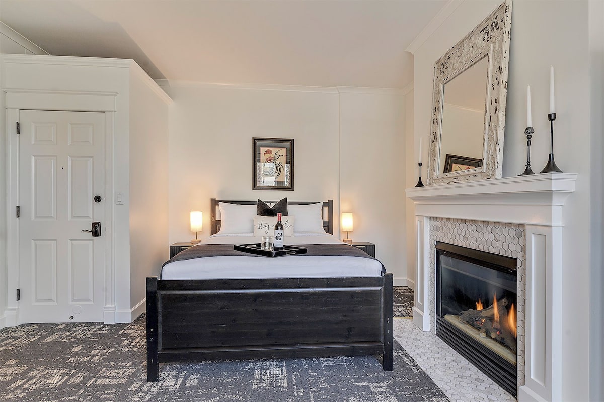 A bedroom with a bed, a fireplace, a door, and a framed artwork on the wall.