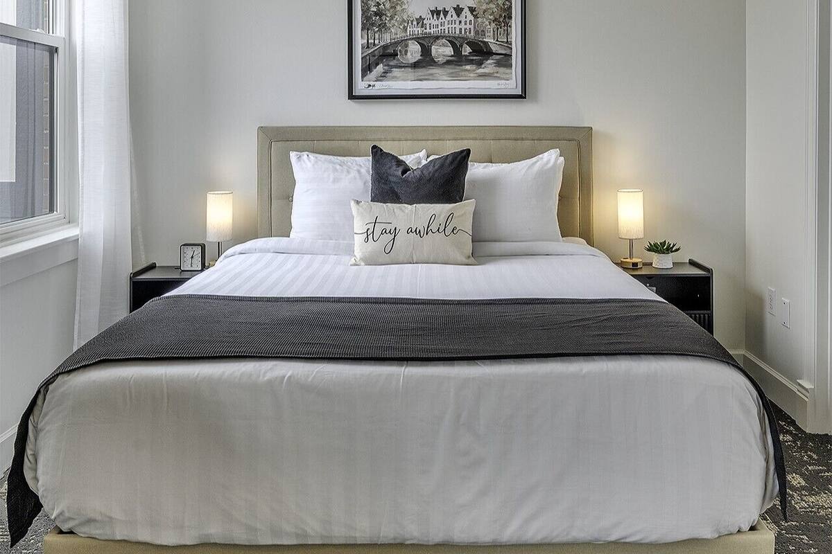A neatly made bed with a headboard, two bedside tables with lamps, a framed picture on the wall, and a window with curtains.