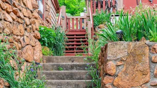 A stone staircase leading to a wooden gate, surrounded by greenery and a red building with a blue and white striped object on the wall. - Surf Shack at Triple Z (Medicine Park)