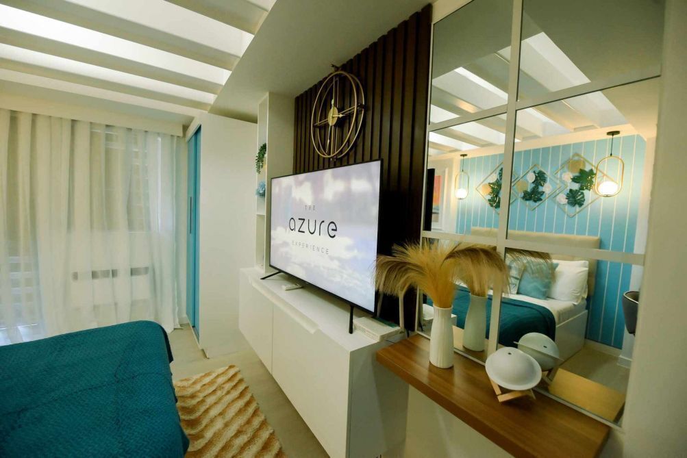 Quarto de hotel moderno com uma grande televisão de tela plana, uma cama com colcha verde-água, uma mesa de cabeceira de madeira e um espelho refletindo o quarto.