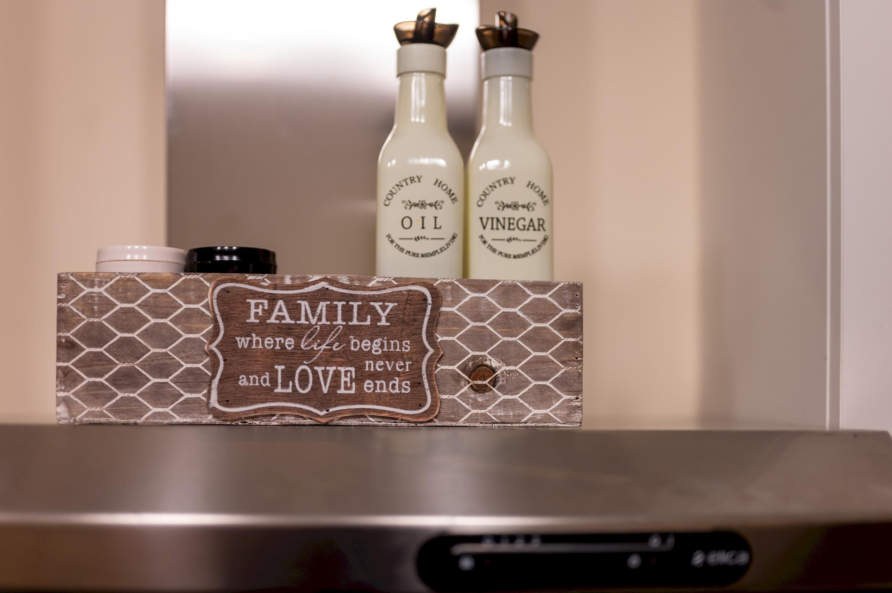 A kitchen counter with a wooden sign that reads 'FAMILY where life begins and LOVE ends,' and two bottles labeled 'COUNTRY HOME OIL' and 'COUNTRY HOME VINEGAR.'