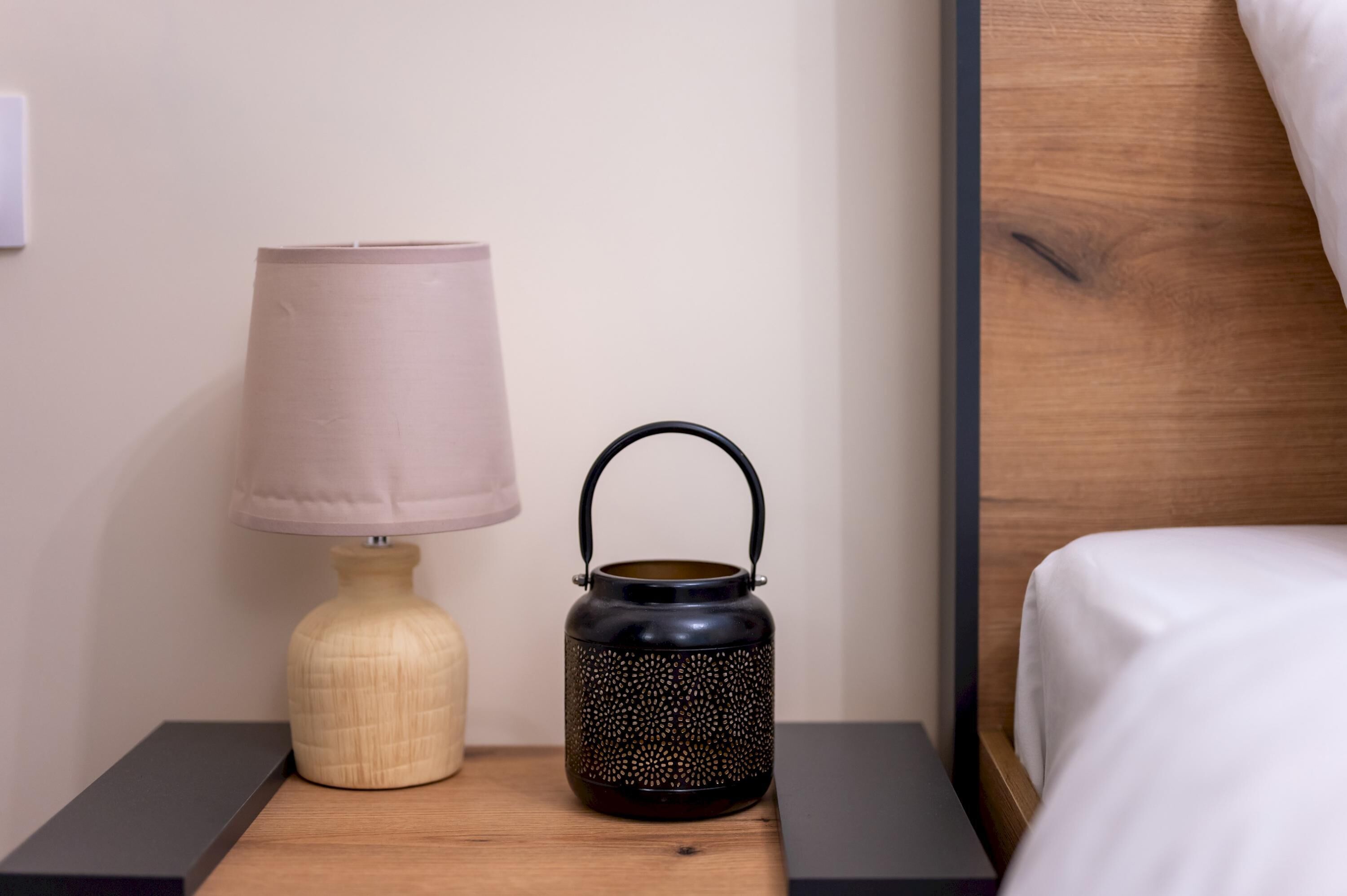 A wooden nightstand with a beige lamp and a black lantern with a handle.