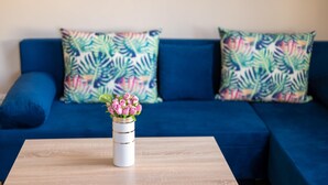 A blue sofa with tropical leaf-patterned pillows and a white vase with pink flowers on a wooden table. - Design Loft in the City Center (Sofia)