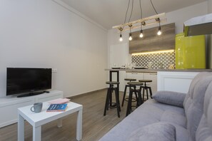 A modern living room with a grey sofa, a white coffee table with books and a mug, a flat-screen TV, and a kitchen area with bar stools and a yellow refrigerator. - Sunny and Modern 1bdr Apartment in the Center (Sofia)