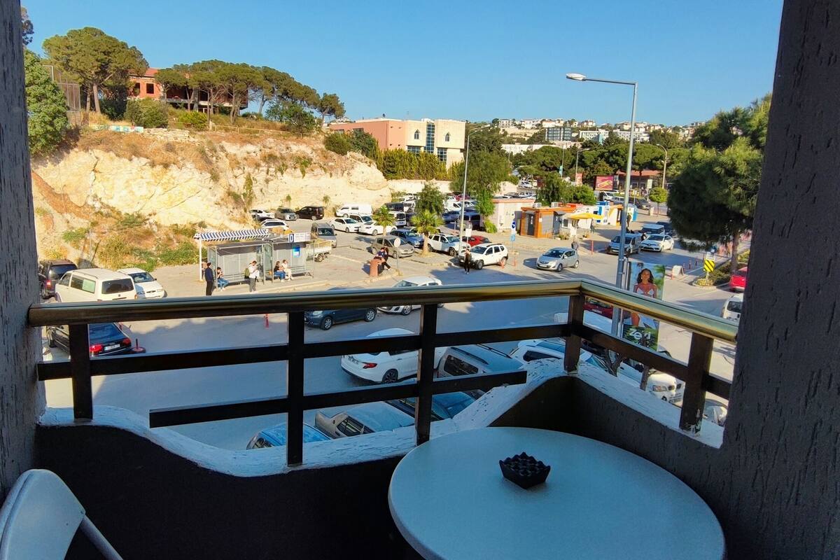 Un balcon avec une table et des chaises, donnant sur une rue animée bordée de voitures garées et d’immeubles.
