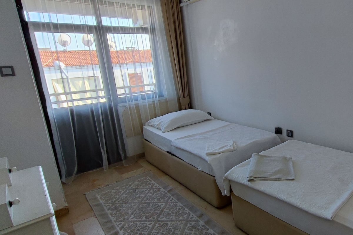 Une petite chambre propre, avec deux lits simples, une fenêtre avec des rideaux et un tapis à motifs sur le sol.