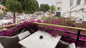 Eine einladende Terrasse mit weißen Tischen und Stühlen, umgeben von leuchtend violetten Blumen und Blick auf eine Straße mit parkenden Autos und Häusern.