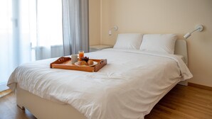 Un letto rifatto con lenzuola bianche, un vassoio di legno con croissant e un bicchiere di succo d'arancia, e un comodino bianco con una lampada.